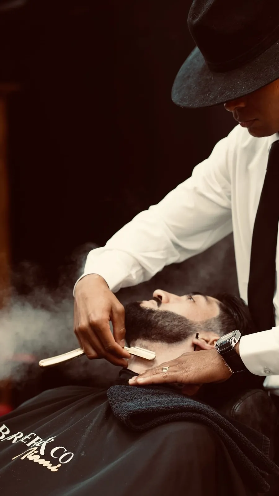 Barber in a fedora giving a straight-razor shave at Barber & Co Miami