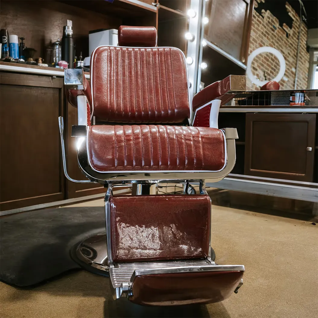 A vintage-style red leather barber chair with chrome accents sits centered in a barbershop, surrounded by wood cabinetry and grooming tools.