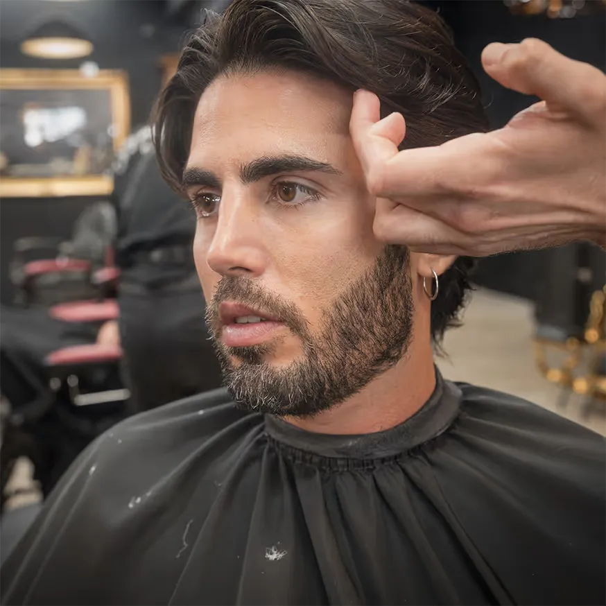 A man with neatly styled, medium-length dark hair and a well-groomed beard sits in a barber chair. A barber’s hand is gently guiding his face to the side, showcasing the clean line-up and detailed beard trim. The barbershop setting features black walls and red chairs in the background.