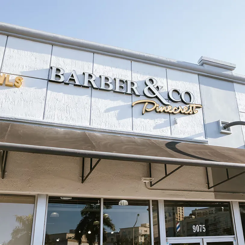 The exterior of Barber & Co Pinecrest, showing a clean white facade with black-and-gold signage and glass windows reflecting palm trees.