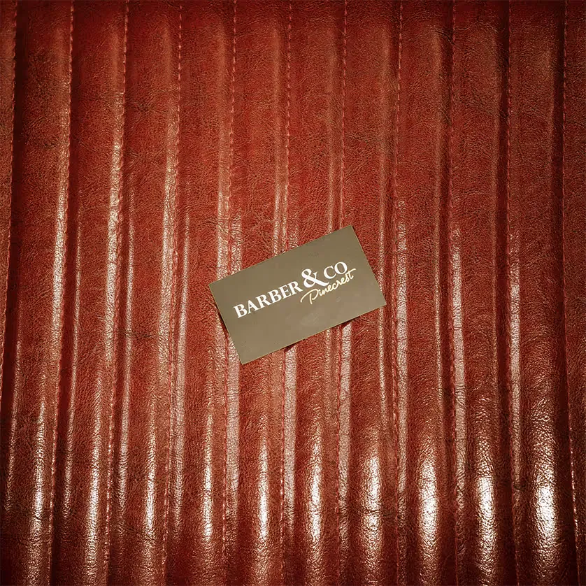 A dark Barber & Co Pinecrest business card resting on a rich, vintage red leather barber chair cushion with vertical stitching.